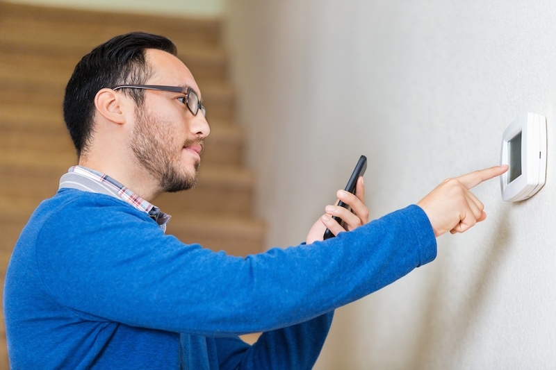 Mid adult hispanic man looks at smart phone for instruction as he learns to use thermostat in his new home. He is touching the digital termostat with his finger. He is standing in front of the stairs leading to the second floor of his home. He is wearing a blue sweater and glasses and has brown hair and a beard.